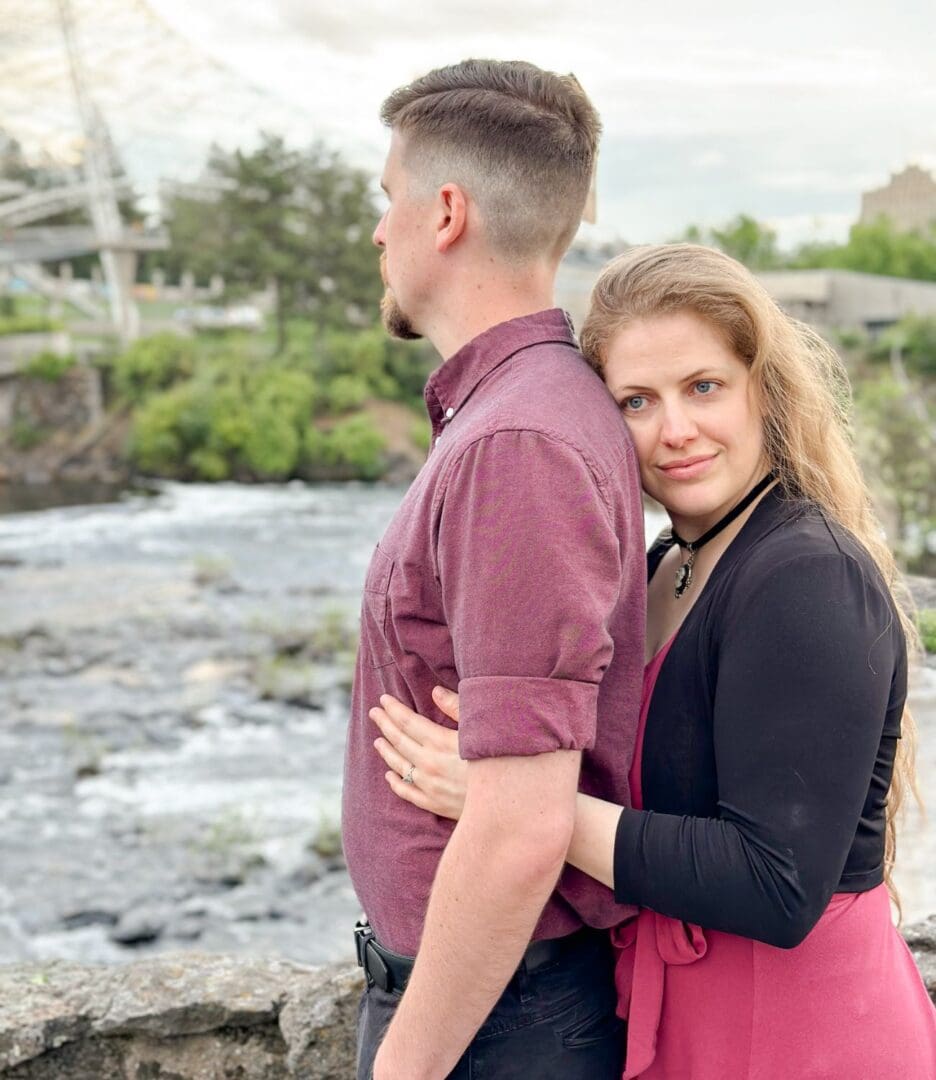 A couple embraces by a river with greenery in the background.