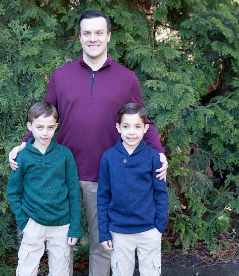 A man stands outdoors with two young boys in front of greenery.