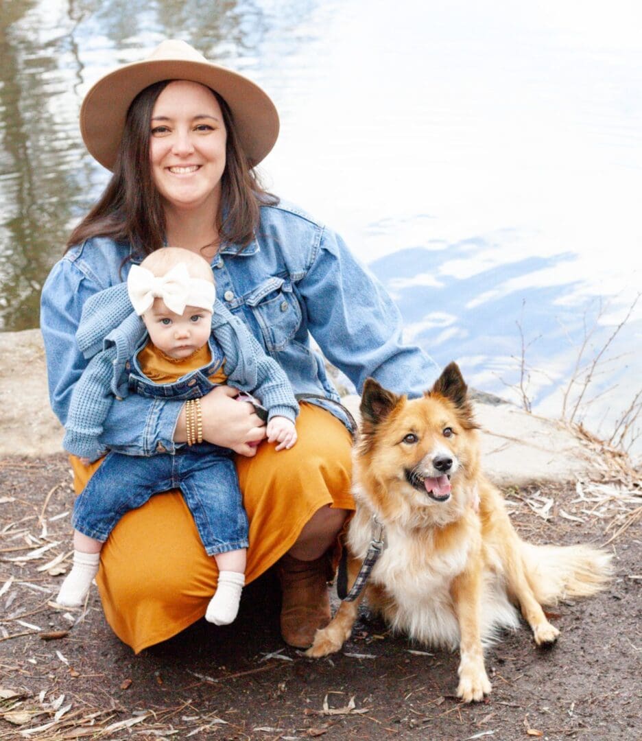 A woman in a hat holding a small dog, sitting beside a larger dog outdoors.