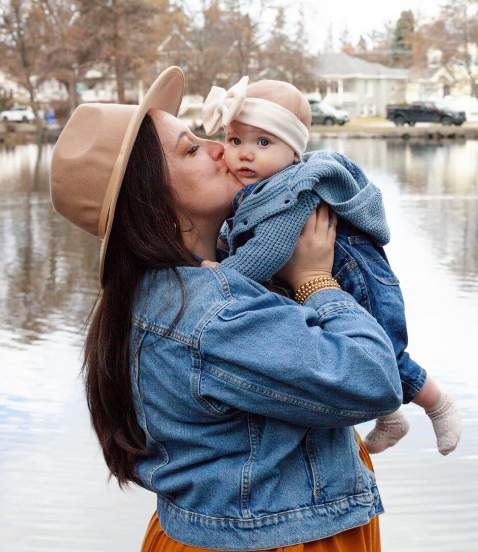 Woman in hat kisses baby by a calm riverside.