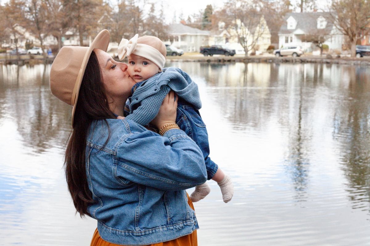 Mother lovingly kisses her baby by a lakeside on a chilly day.