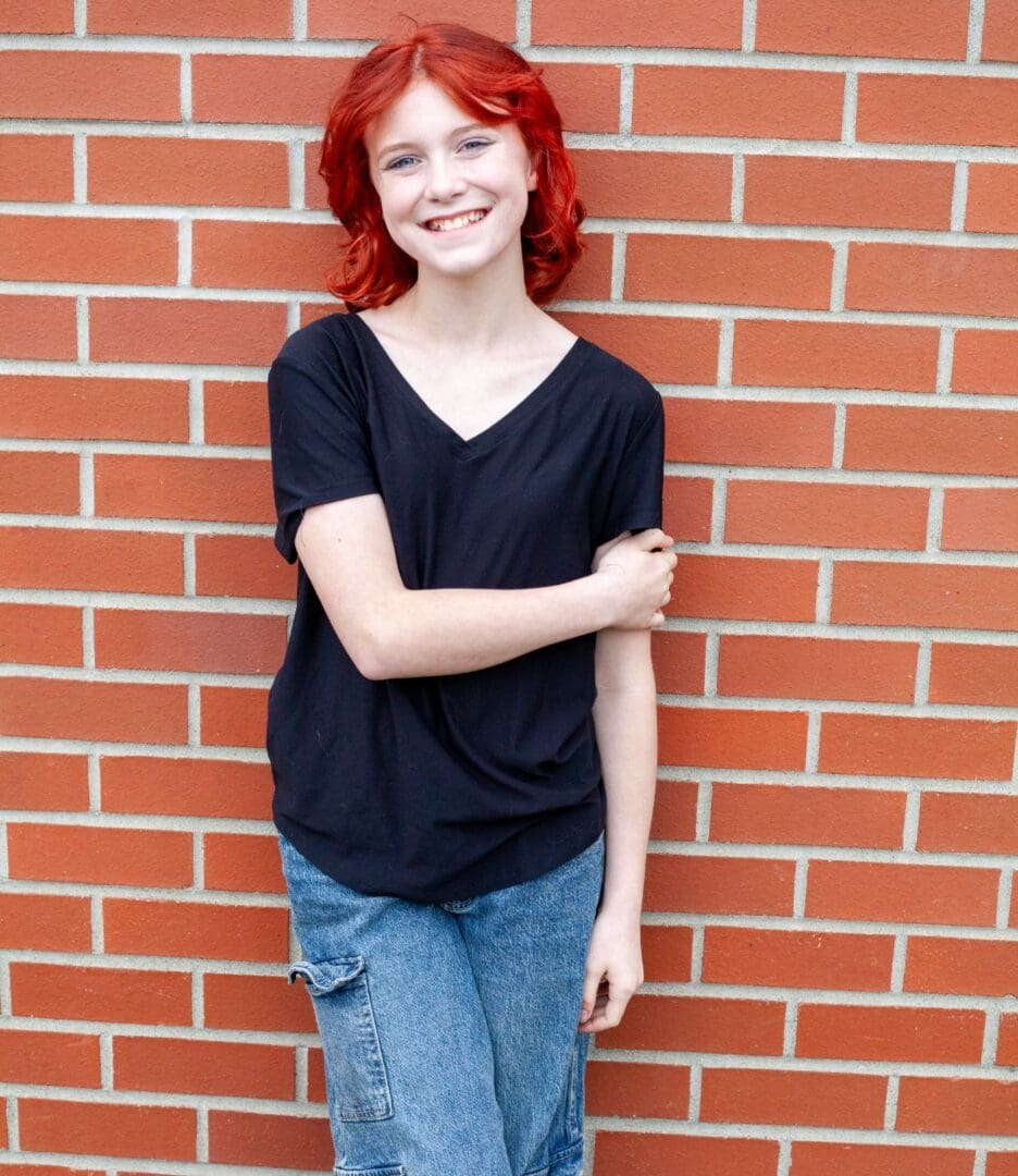 Smiling young person with red hair standing against a brick wall.