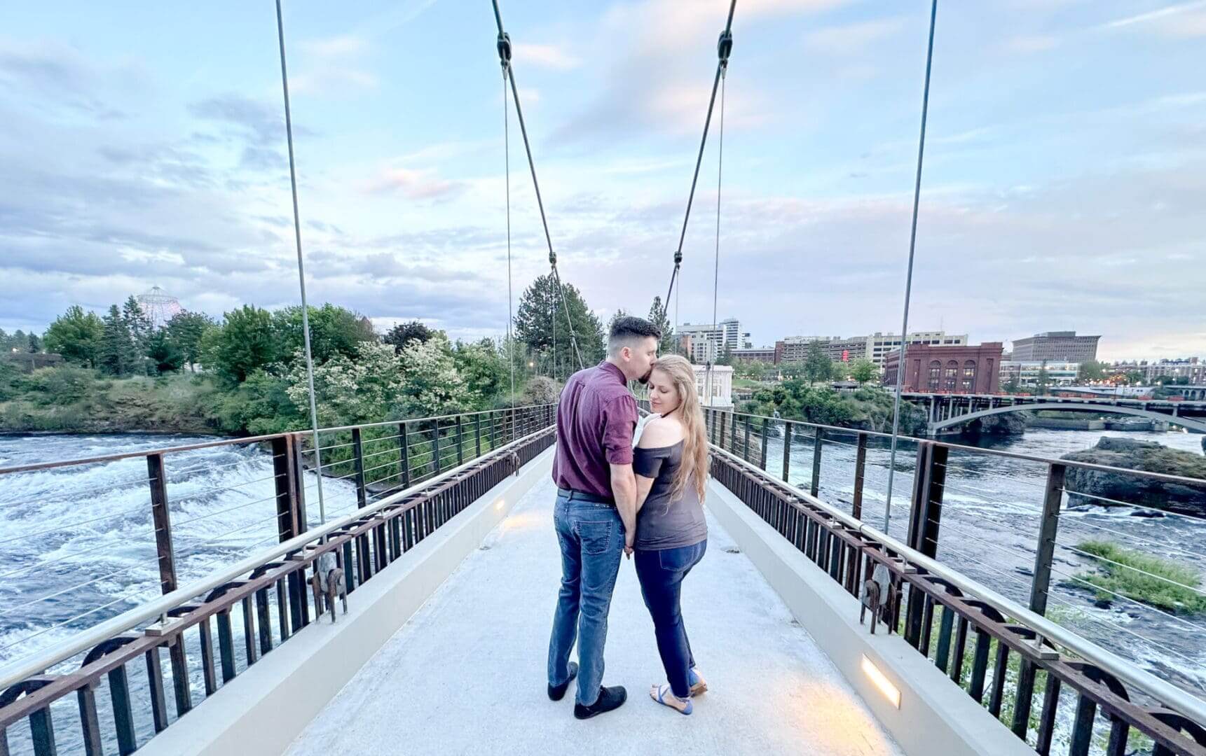 Couple sharing a tender moment on a scenic suspension bridge under a blue sky.