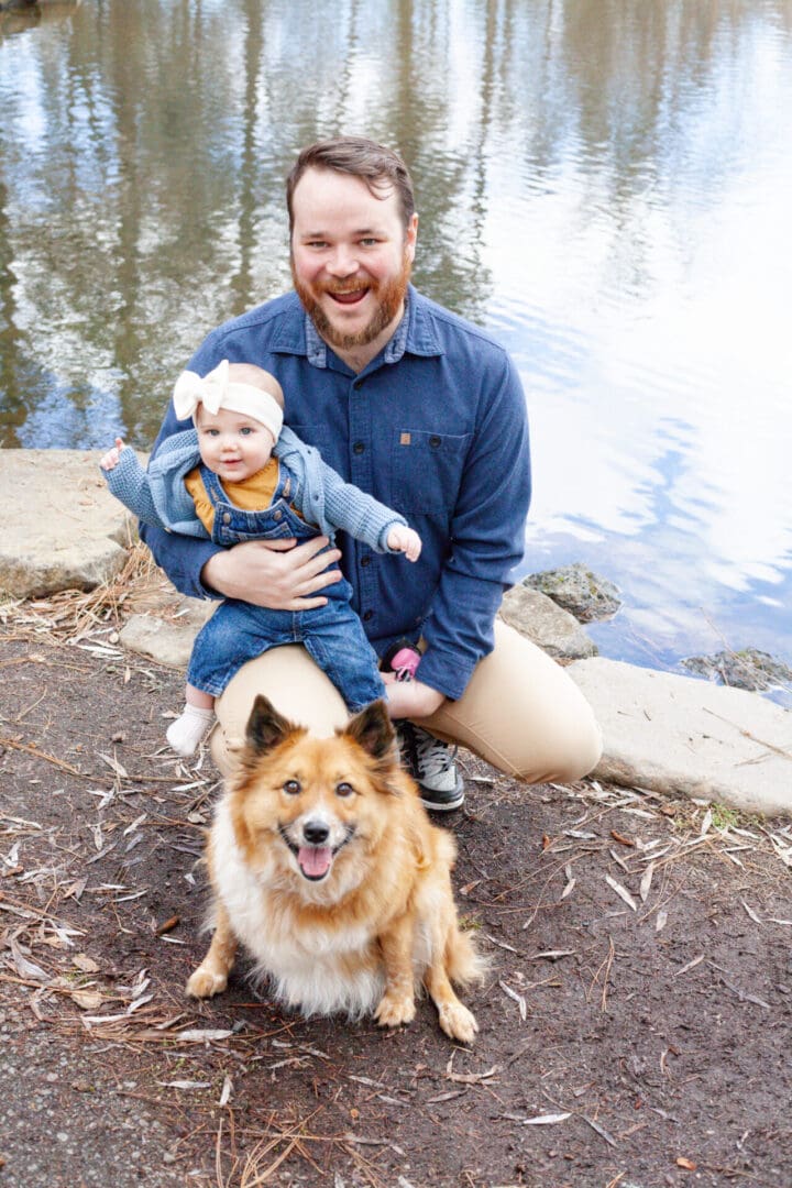 Man kneeling by a lake holding a baby with a fluffy dog in front.