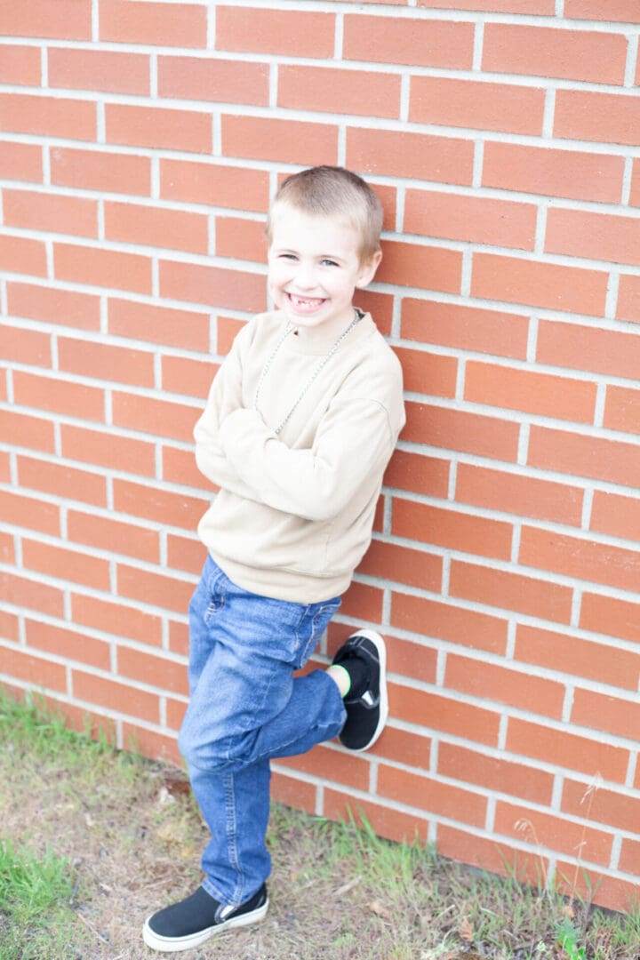 A smiling boy leaning against a brick wall with crossed arms.