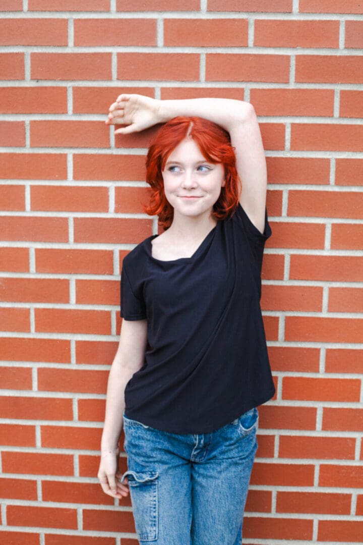 Smiling woman with red hair standing against a brick wall, arm raised.