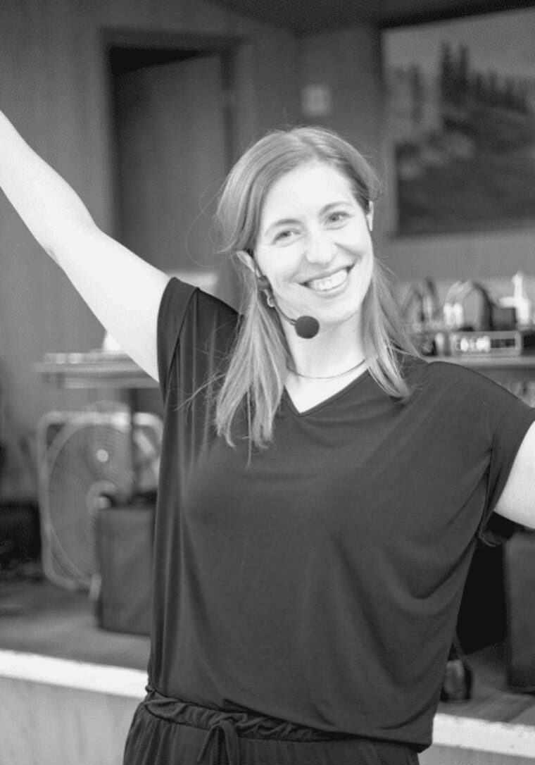 A smiling woman with a headset microphone, raising one arm.